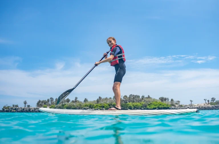 Person wearing a red life vest stands on a paddleboard, paddling in bright turquoise water under a clear blue sky.