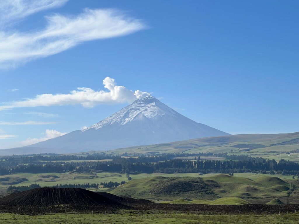 Snow-covered volcanic mountain rising above green rolling hills under a clear blue sky with scattered clouds in the distance.