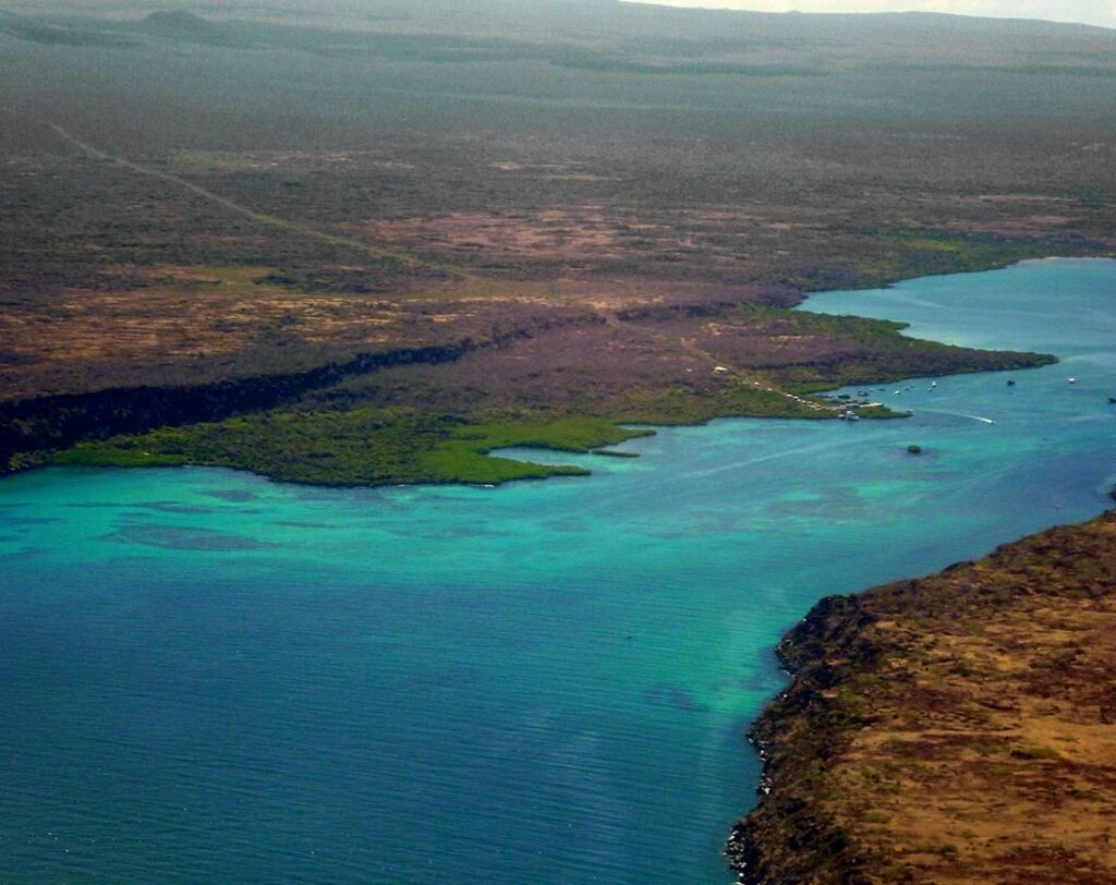 Aerial view of a turquoise bay along a rugged coastline with dark volcanic rocks and a few boats nearby plunge of water.