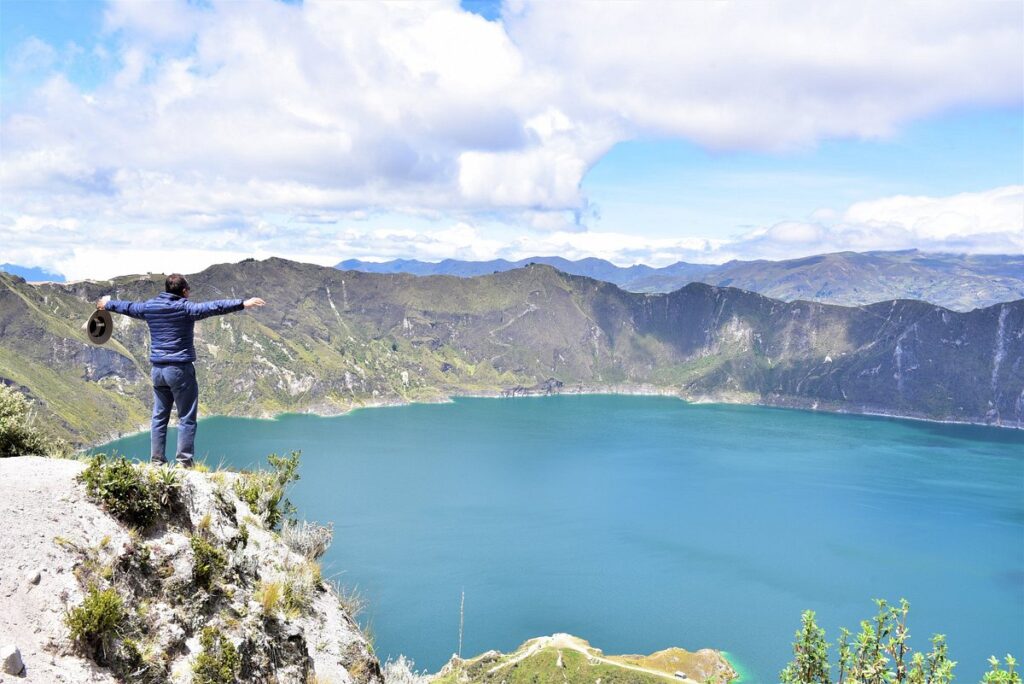 Person standing on a rocky cliff with arms outstretched, holding a hat, overlooking a turquoise crater lake and mountains.