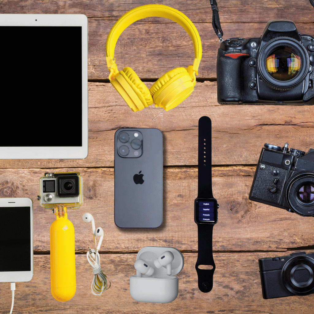 Top-down view of gadgets on a wooden table: tablet, yellow headphones, DSLR camera, iPhone, Apple Watch, AirPods case, and a GoPro with yellow housing.