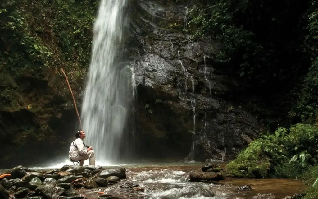 Man in light clothing sits on a rock beside a tall waterfall in a lush, forested setting. Wide, wet rocks and a shallow pool at the base are visible.