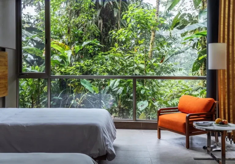 Hotel room with a large glass wall overlooking dense tropical greenery; orange armchair and a small table with fruit beside a white bed(s).