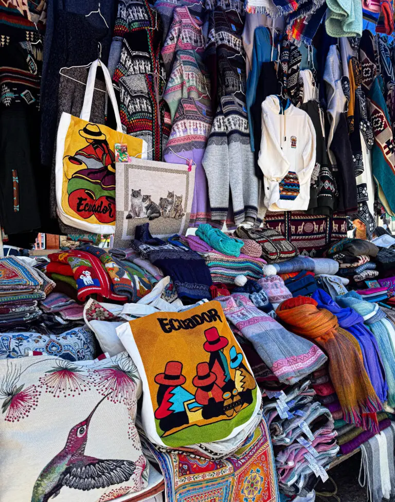 Colorful market stall with woven scarves, sweaters, and bags; front includes bright Ecuador-branded tote bags.