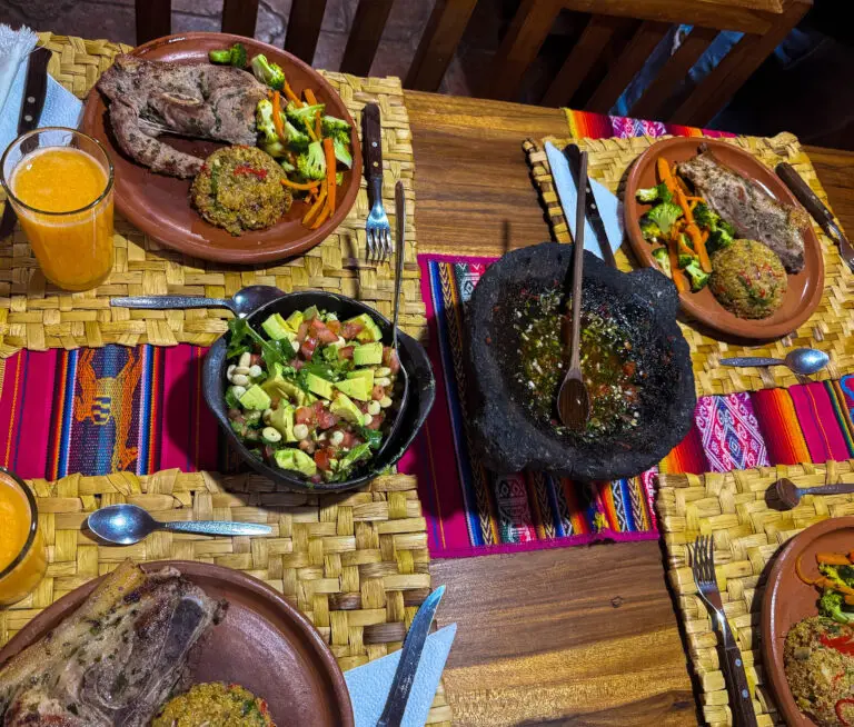 Table with two clay plates of roasted meat, vegetables, and salad on a woven mat, plus a glass of orange juice and colorful textiles nearby.