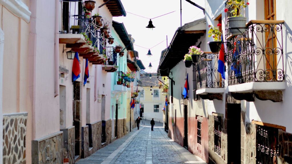 Narrow cobblestone street lined with pastel buildings, balconies, hanging plants, and flags; a sunlit alley with a few distant pedestrians.00Narrow Mediterranean alley with wrought-iron balconies, potted plants, and blue-red flags along a cobblestone street.