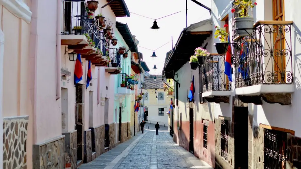 Narrow cobblestone street lined with pastel buildings, balconies, hanging plants, and flags; a sunlit alley with a few distant pedestrians.00Narrow Mediterranean alley with wrought-iron balconies, potted plants, and blue-red flags along a cobblestone street.