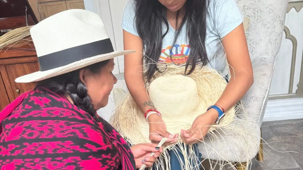 Two women work together to weave a straw hat outdoors, inspecting the straw strands as one guides the other.