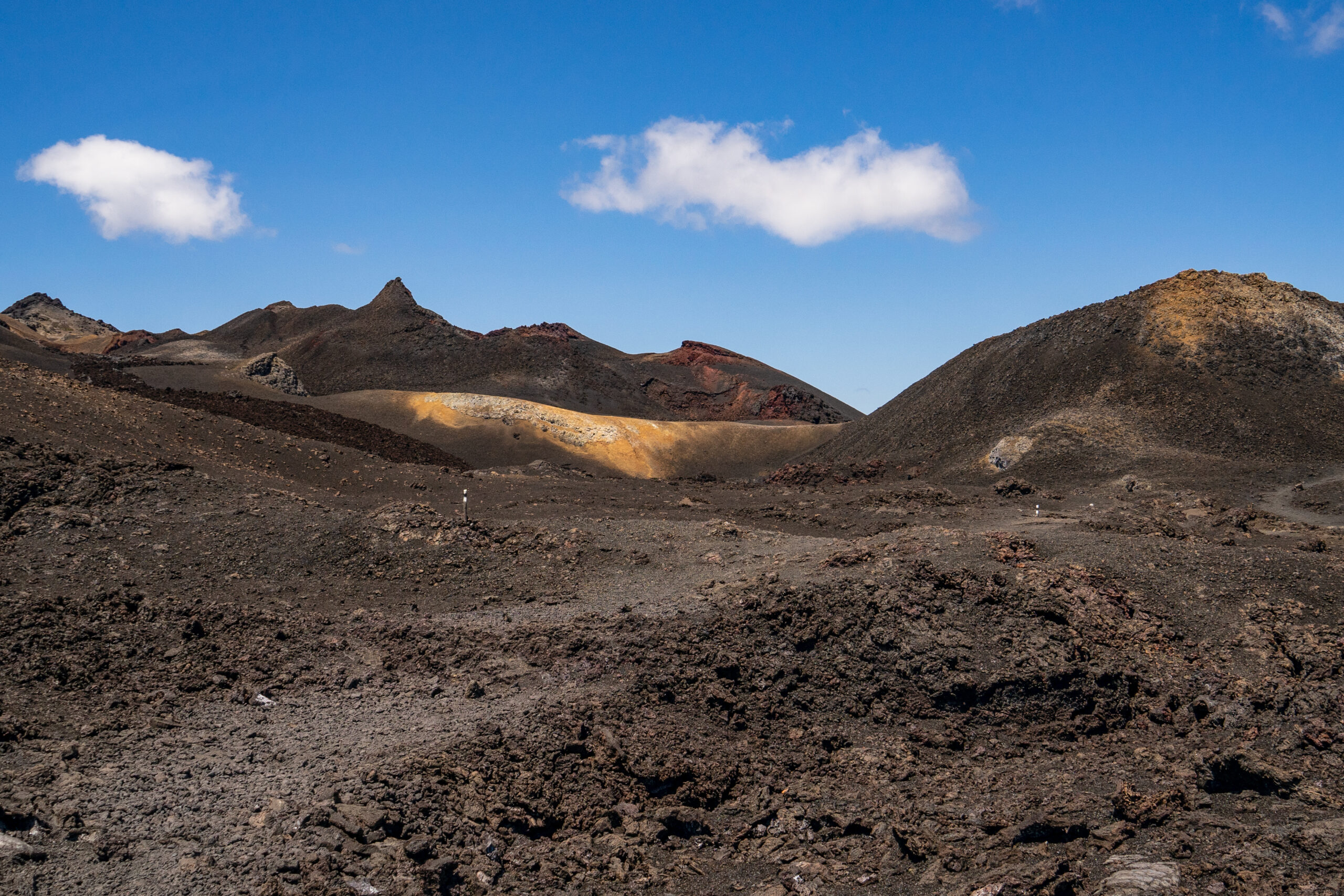Barren volcanic terrain with dark rocky mounds against a bright blue sky and a few white clouds.