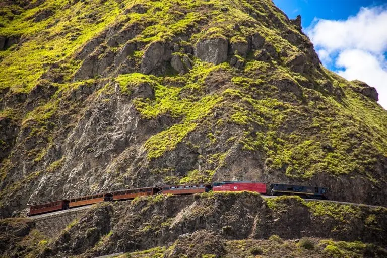 Red locomotive pulls a line of brown passenger cars along a cliffside railway carved into a rocky, green hillside under a blue sky.