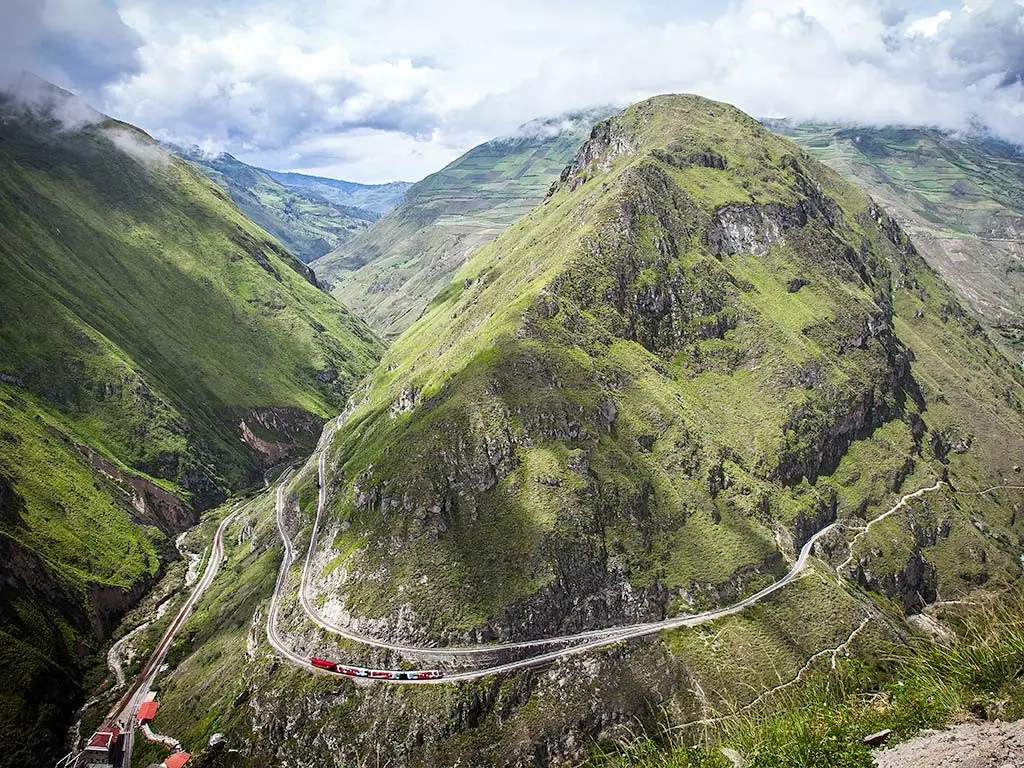 Winding mountain road with sharp switchbacks along a green cliff, a red train car visible on the curves below.