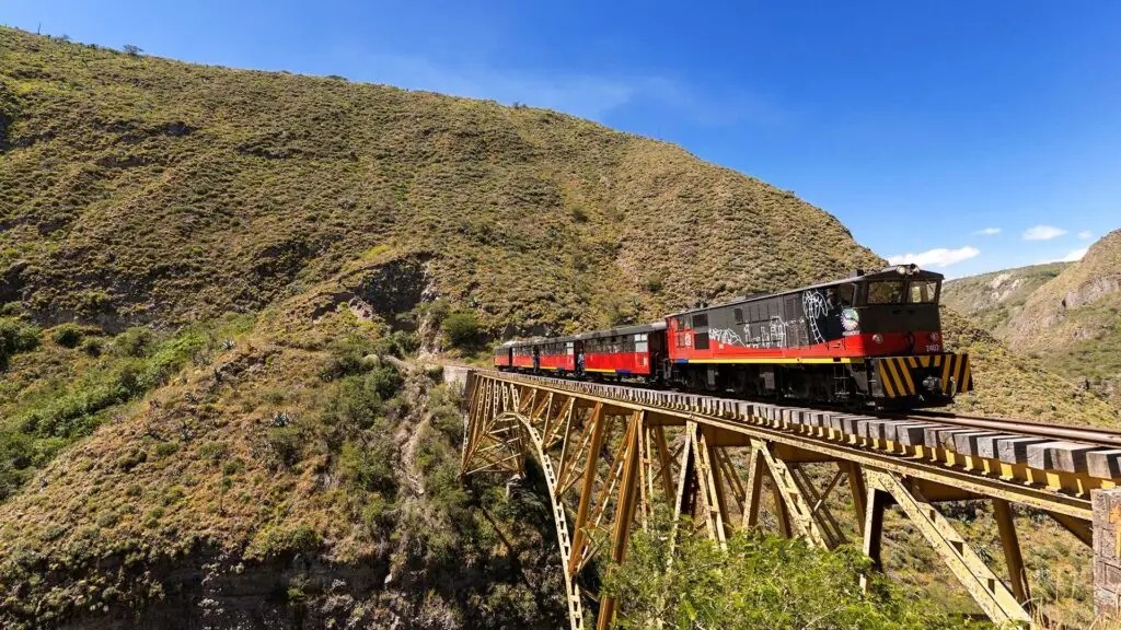 Diesel locomotive and red passenger cars cross a tall steel bridge over a rocky canyon with brown hills. (Informative)