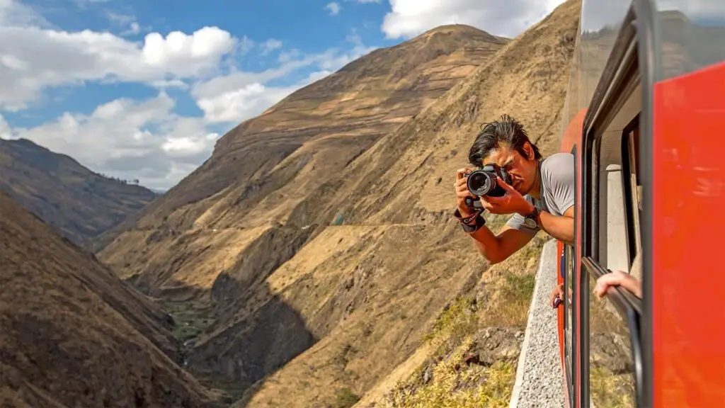 Photographer leaning out of a red train window, aiming a DSLR at rugged canyon scenery below