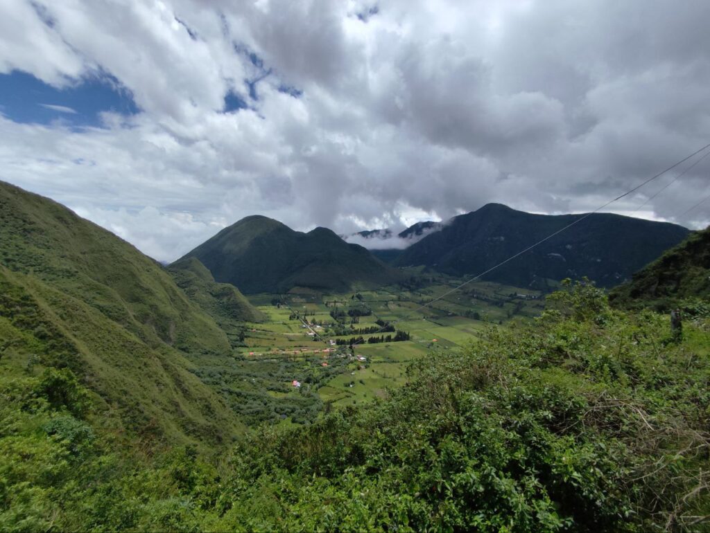 Green valley between rounded, forested mountains under a cloudy sky, with a small village and patchwork fields in view.