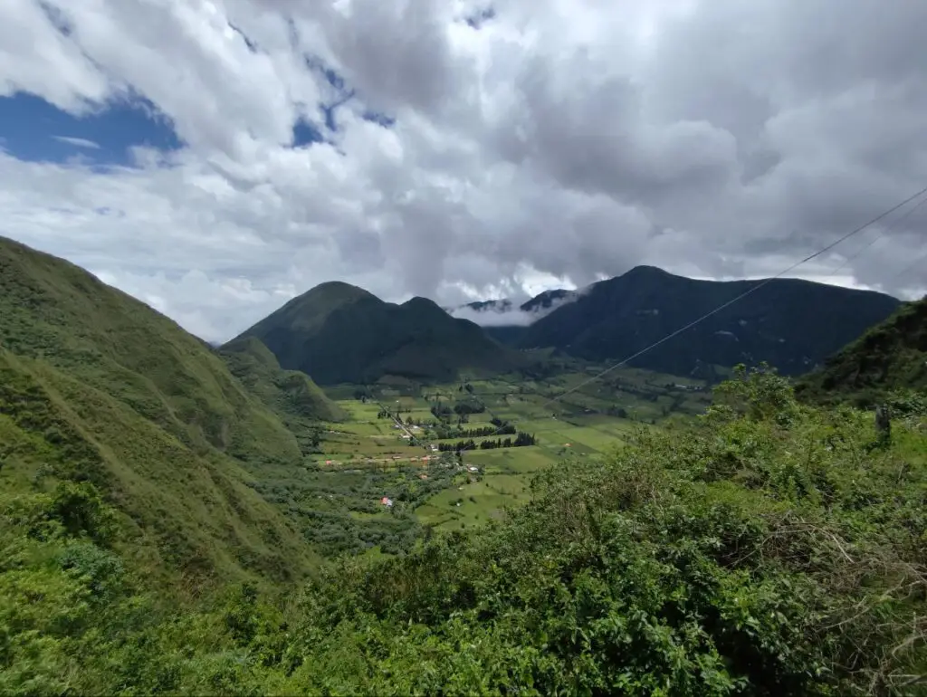 Green valley between rounded, forested mountains under a cloudy sky, with a small village and patchwork fields in view.