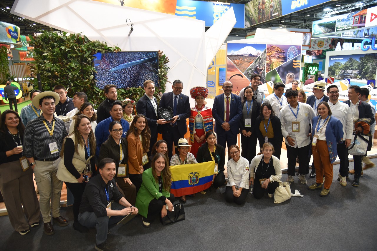 Group of diverse people posing for a photo at a trade show booth, holding an Ecuador flag, with some in traditional dress and name badges visible.