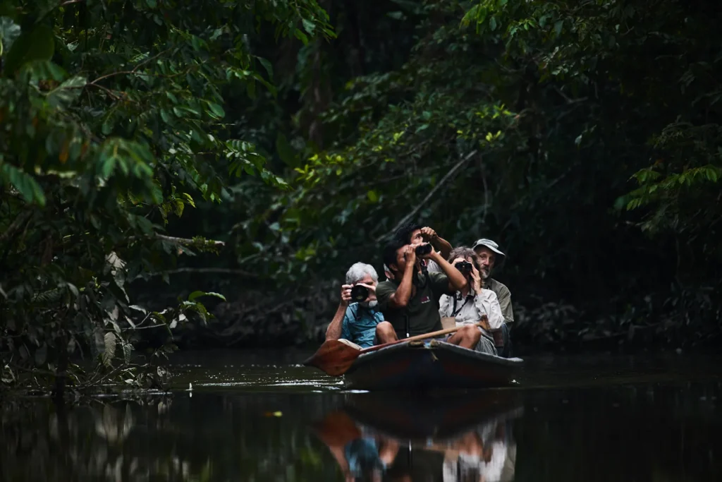 Five people in a small boat on a dark river, surrounded by dense green jungle, some using cameras to photograph wildlife.