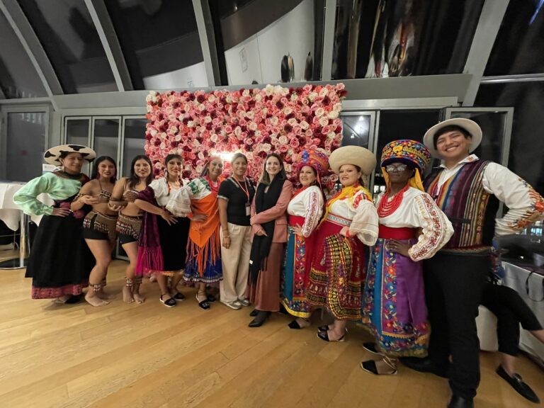 Group of people in colorful traditional costumes posing together in front of a pink rose backdrop, smiling at the camera.