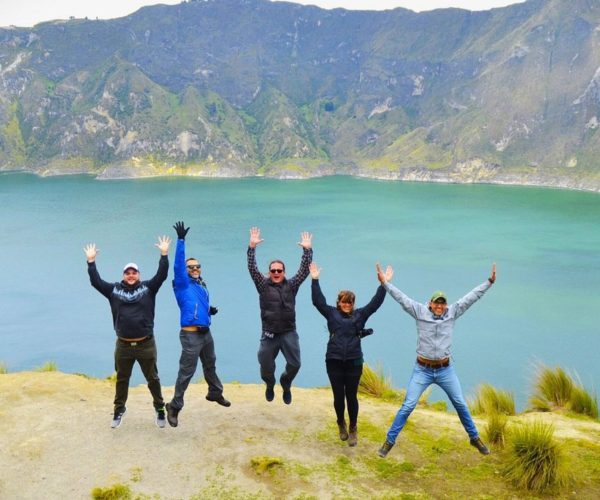 Six people jumping in the air with arms raised at a rocky lakeside, turquoise water and green mountains in the background.