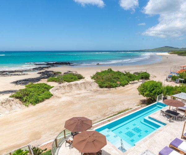Resort beach view with turquoise sea, white sand, rocky patches, and a poolside area with lounge chairs and umbrellas in the foreground.