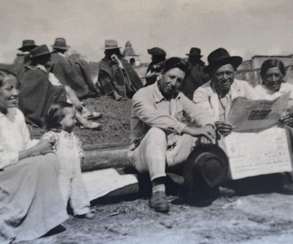 Black-and-white photo of a family sitting on a low wall outdoors; a braided girl sits left, several adults with hats in the middle, and one person reads a newspaper at right.