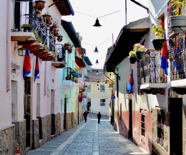 Narrow cobblestone street lined with pastel buildings, balconies, hanging plants, and flags; a sunlit alley with a few distant pedestrians.00Narrow Mediterranean alley with wrought-iron balconies, potted plants, and blue-red flags along a cobblestone street.