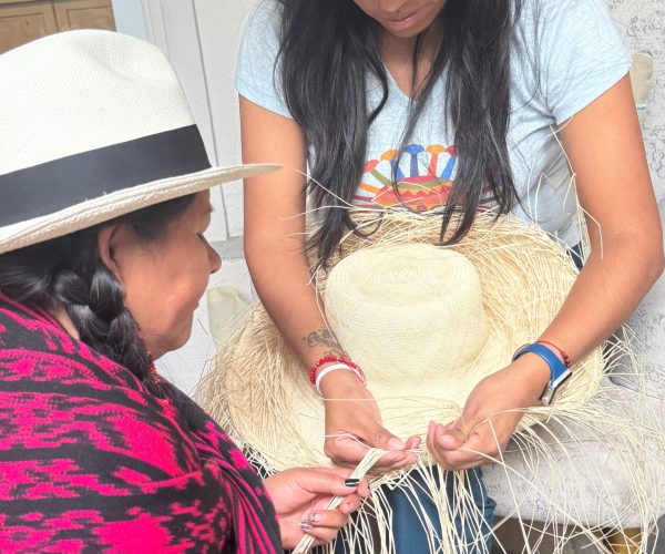 Two women work together to weave a straw hat outdoors, inspecting the straw strands as one guides the other.