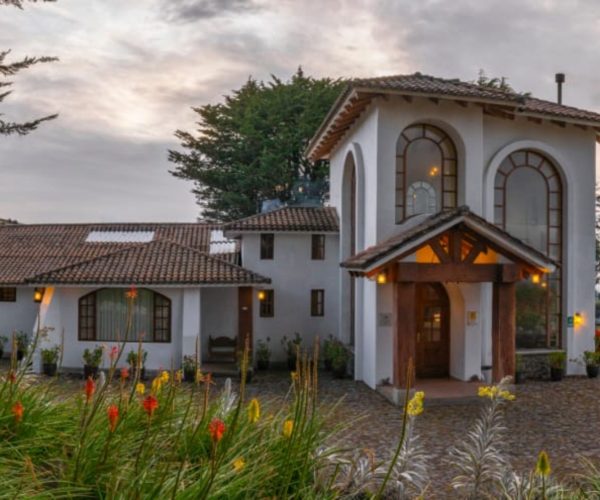 White stucco villa with red tiled roof and tall arched windows, wooden entrance, and a paved courtyard surrounded by flowering plants.